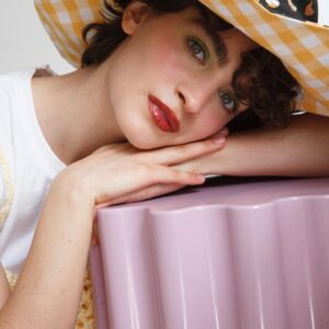 Modelo con sombrero amarillo de cuadros grandes, estilo reversible, llamado Capote margarito, apoyada en un objeto lila, mirandonos con ojos cálidos
